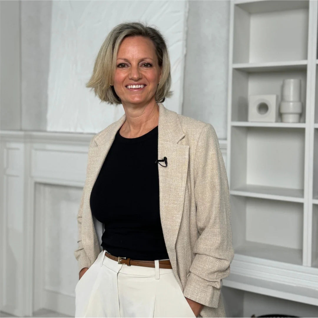 Woman in a beige blazer over a black top and white pants standing in a room with white walls and shelves.