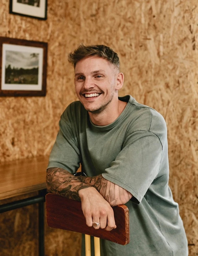 Man with tattoos wearing a green shirt sitting on a wooden stool against a wooden wall.