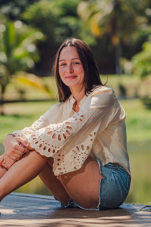 Woman sitting outdoors wearing a white lace top and denim shorts with a blurred natural background