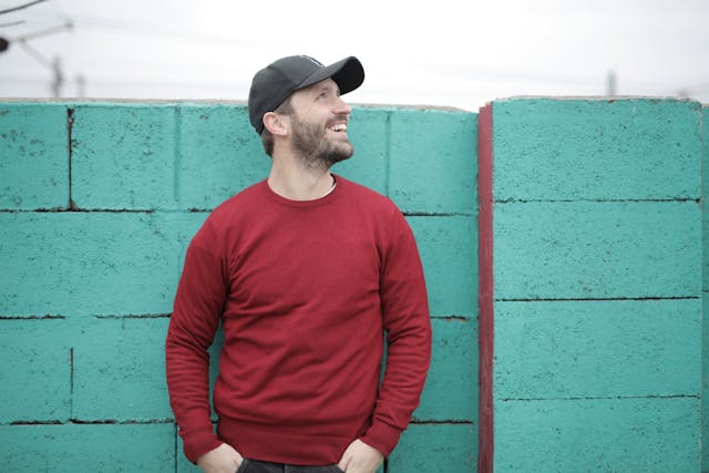 Man wearing a red sweater and black cap standing against a teal brick wall.