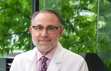 Man in a white coat and glasses standing in front of greenery